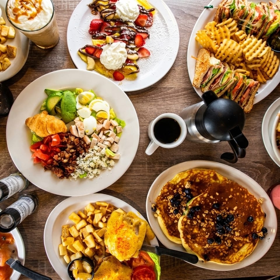 A wooden table laden with various breakfast and brunch dishes, including eggs, bacon, pancakes, and sandwiches.